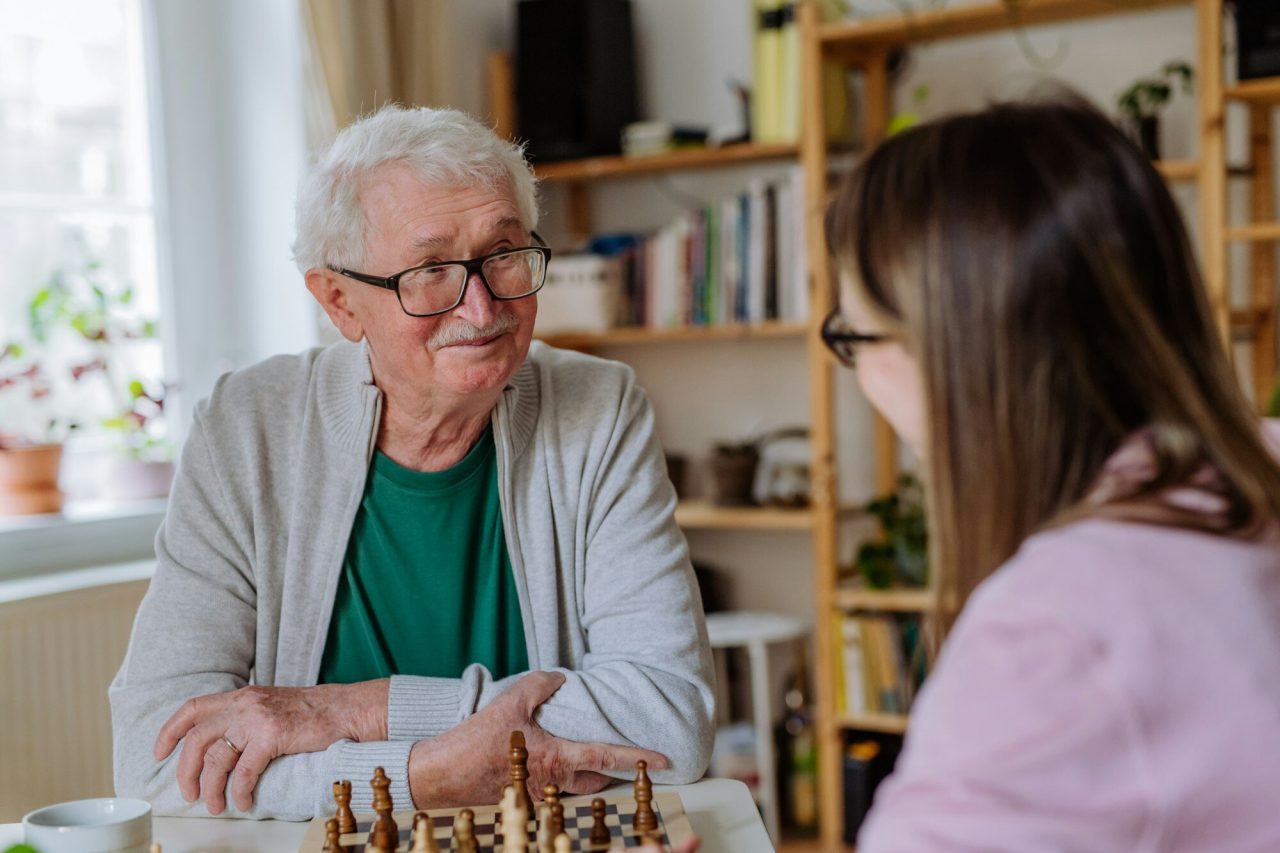 Adult daughter visiting her senior father at home and playing chess together.